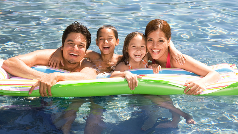 Father, son, daughter, and mother all smiling at the camera while in the pool holding a pool float
