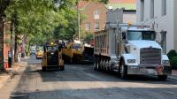 A white truck carrying asphalt and a bobcat vehicle repave an Alexandria street