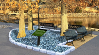 picture of a memorial with a sign, surrounded by trees, four benches a sign and river rocks